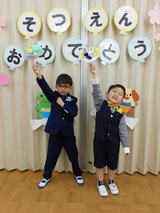 卒園生二人が、「そつえんおめでとう」と書かれた掲示物の前で、片手をあげたポーズをとっている記念写真