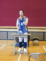 体育館フロアの前方中央で、マイクを片手に持って片岡さんが講演をしている様子